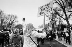 a black and white photo of a group of protestors. One of them holds a sign that ways "we are better than this!"