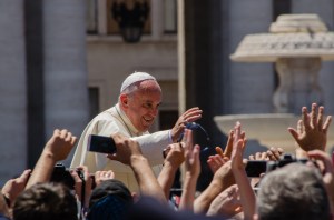 Pope Francis blesses a group of people with raised hands, some holding cameras and phones, in front of a stone fountain.